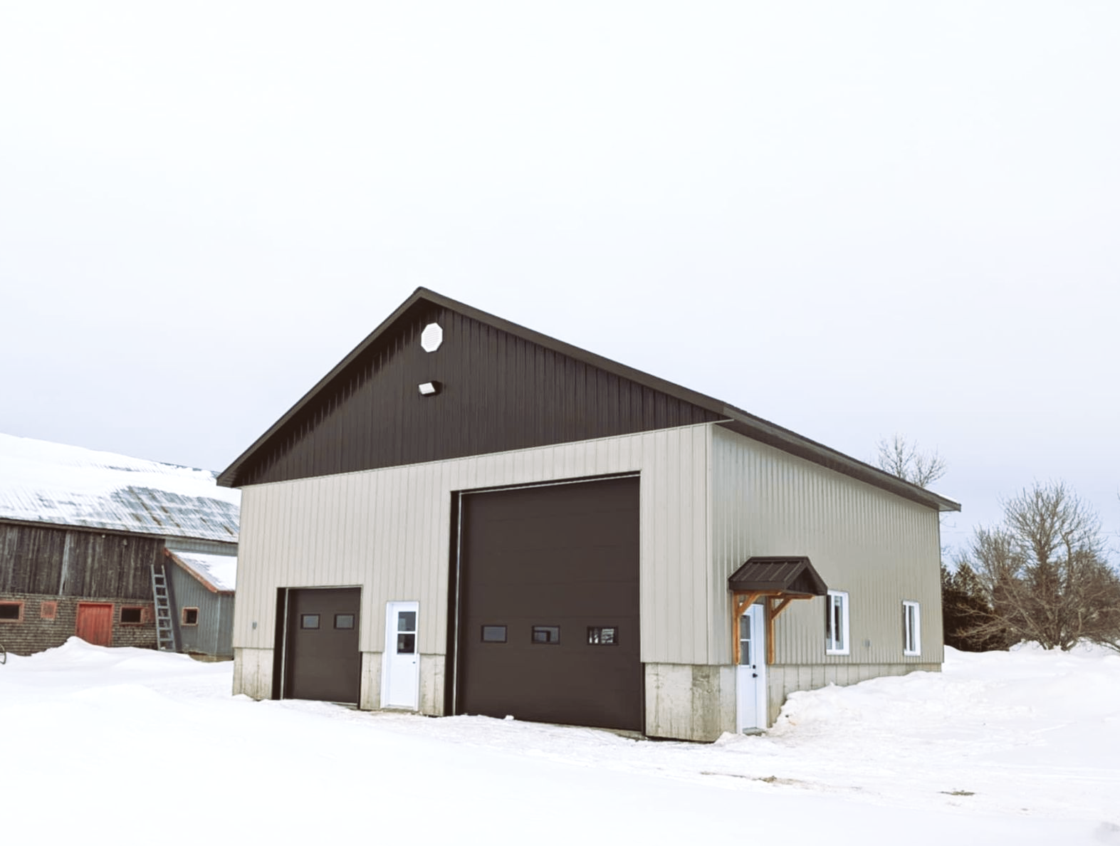 Bâtiment métallique de garage ou d'atelier recouvert de neige, avec un toit brun foncé et de grandes portes sectionnelles.