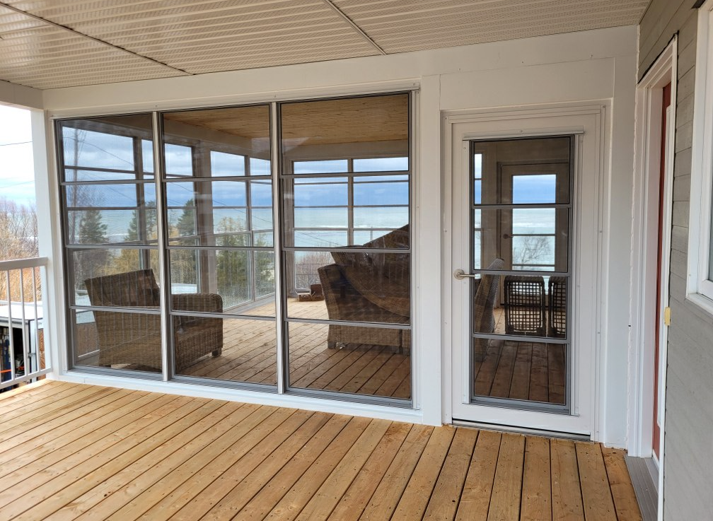 Véranda avec plancher en bois, portes coulissantes en verre et porte latérale blanche donnant sur une vue ensoleillée à l'extérieur.