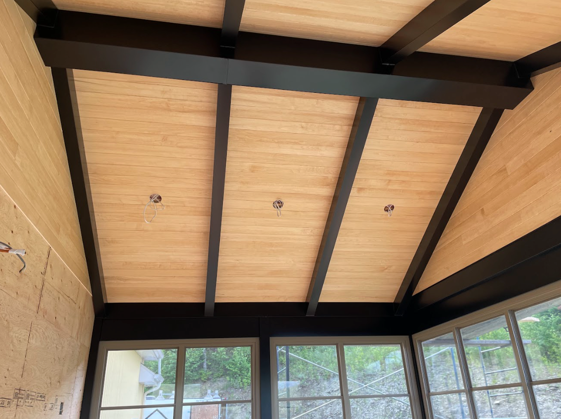 Plafond voûté lambrissé avec poutres noires au-dessus d'une véranda aux grandes fenêtres.