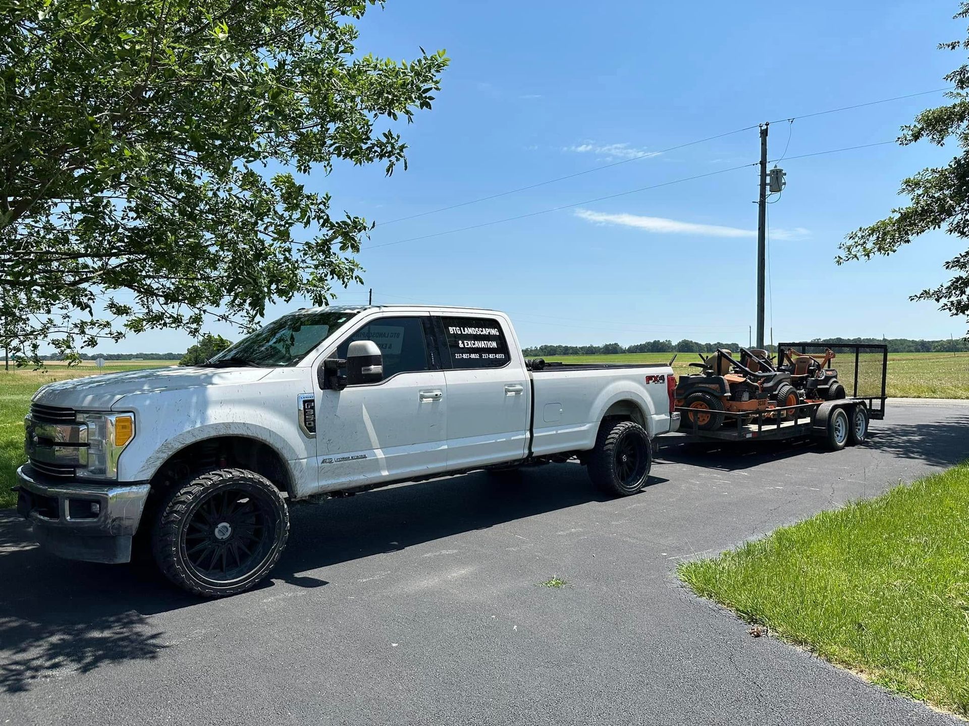Silver pickup truck towing a trailer with landscaping equipment on a sunny day.
