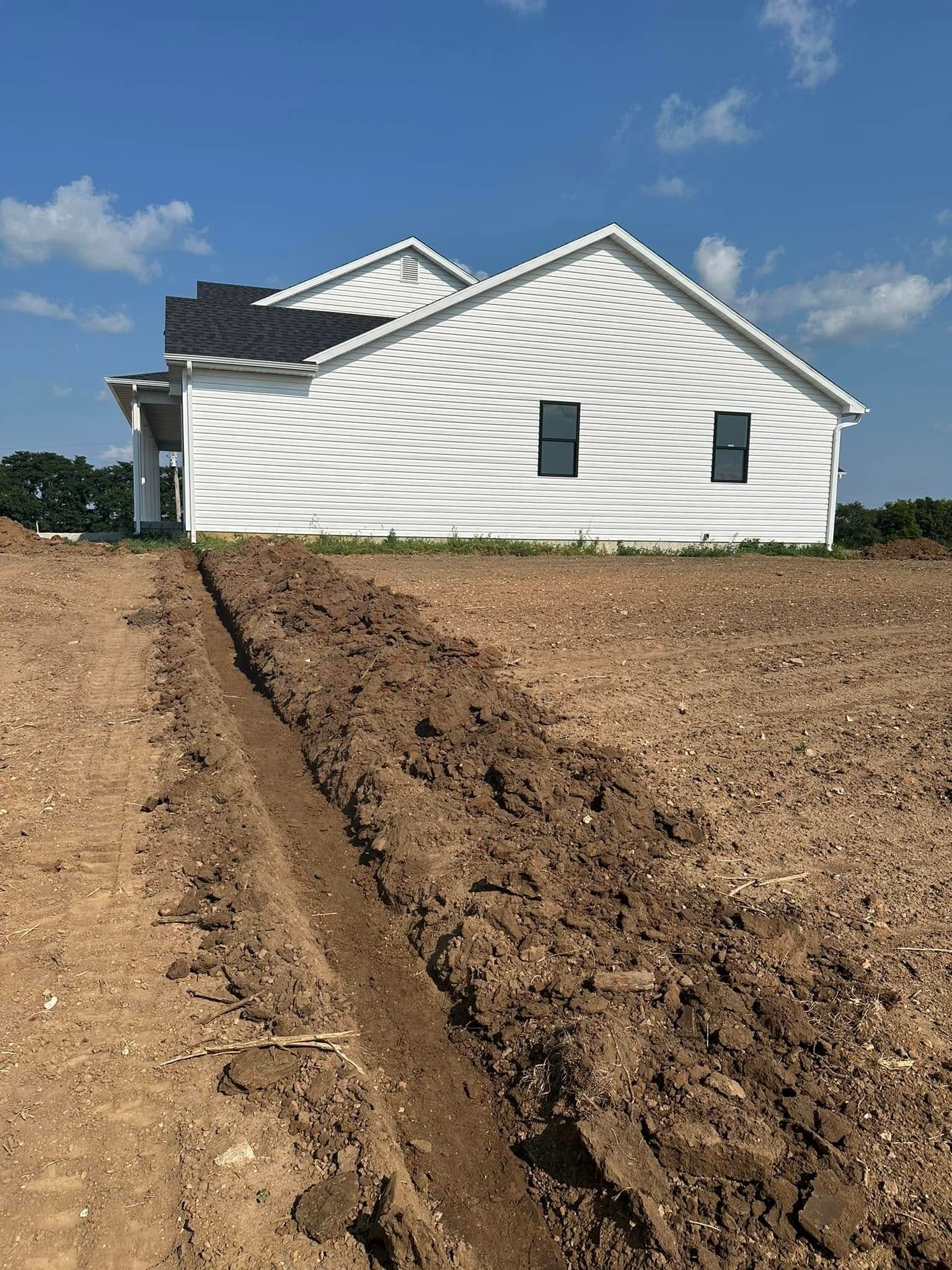 A newly built white house with dark trim and roof; a trench in the foreground.