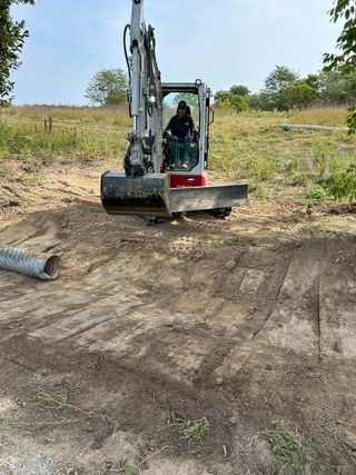 An excavator digs into dirt. A person is operating the machine on a grassy area.