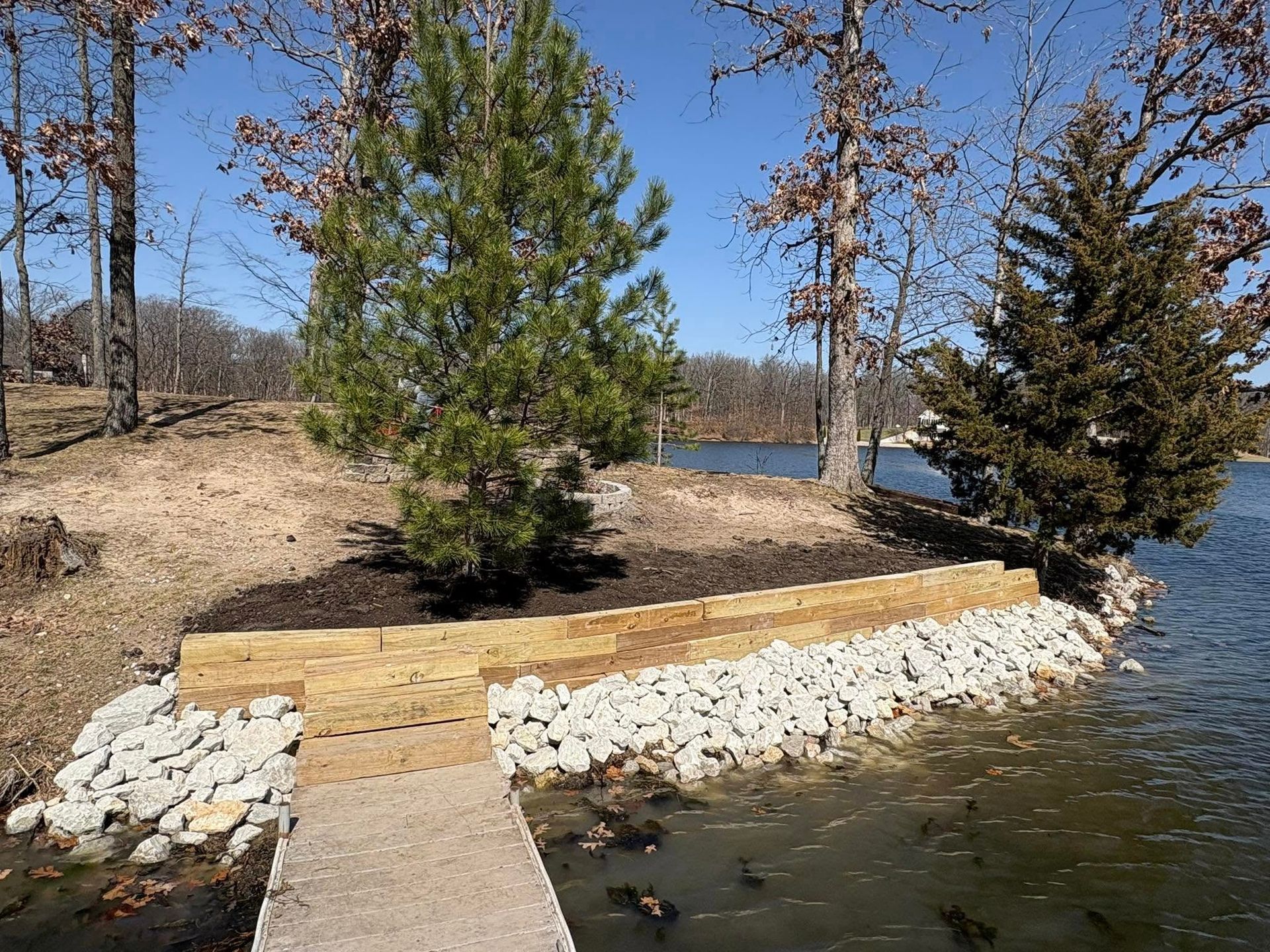 Lakeside view with a wooden retaining wall, rocks, and trees under a blue sky.