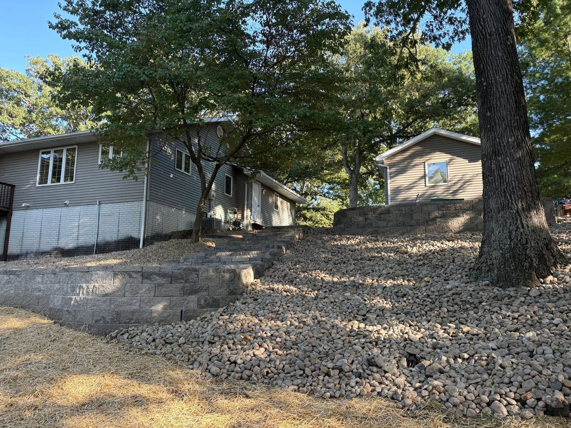House and outbuilding on a sloped lot covered in gravel, with retaining walls.