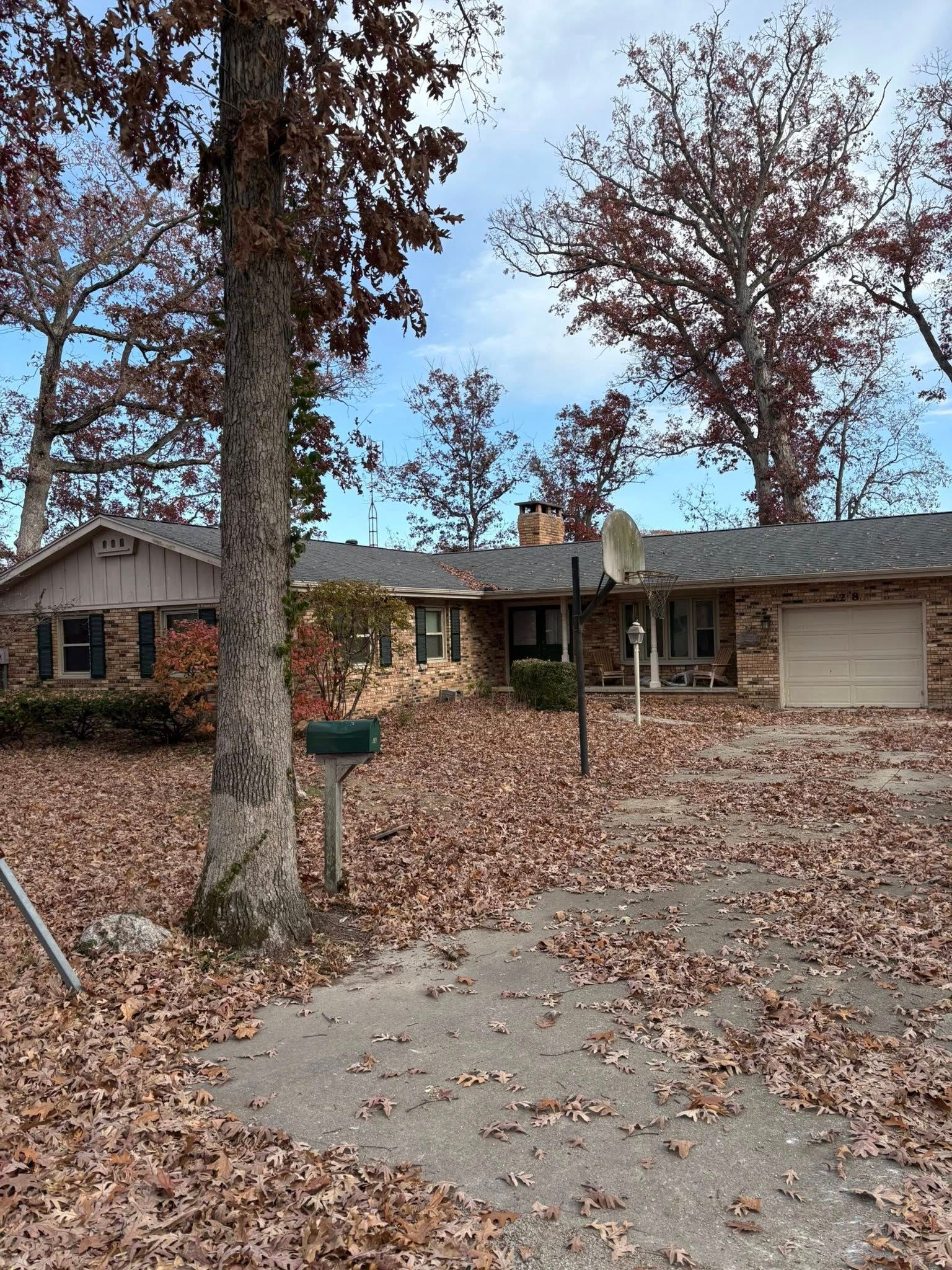 A one-story brick house with a driveway covered in fall leaves. Trees with red leaves frame the house under a blue sky.