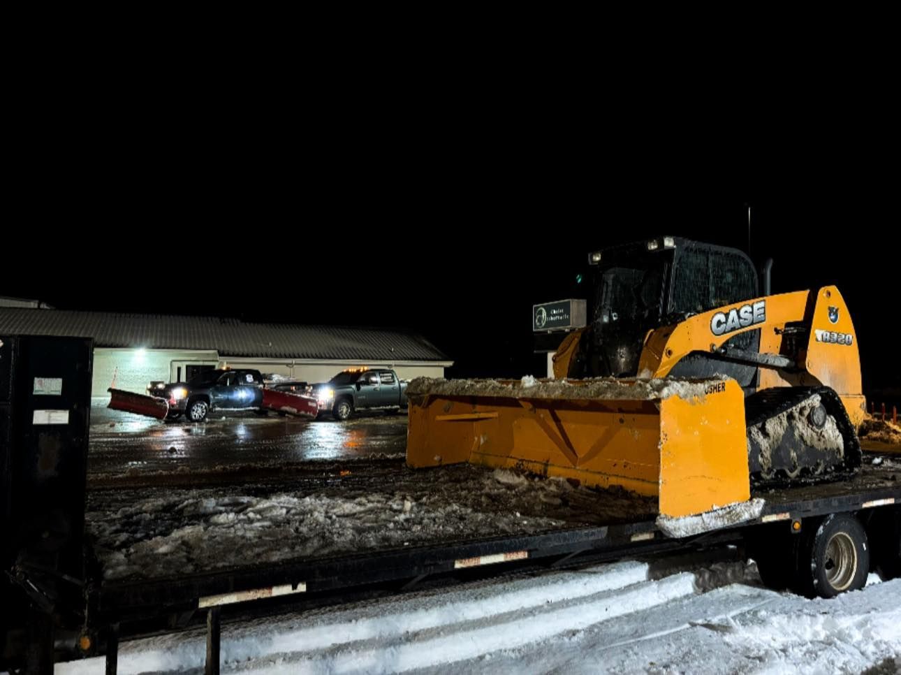 Yellow snowblower on a trailer, parked at night. Building and vehicles are in the background.