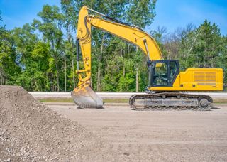 Yellow excavator next to a pile of gravel on a paved surface, trees in the background.