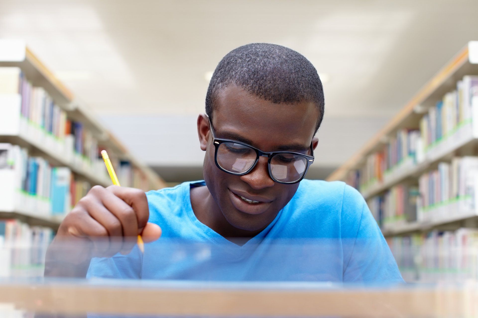 Young person in glasses writing at a desk in a library, surrounded by bookshelves.