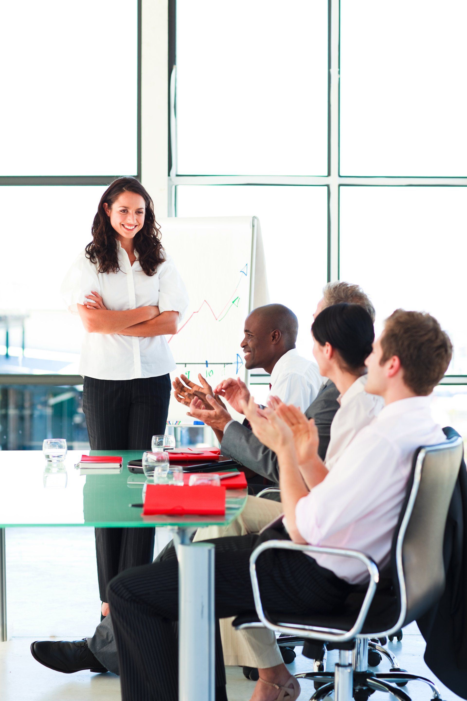 Woman with crossed arms smiles as colleagues clap in a conference room.