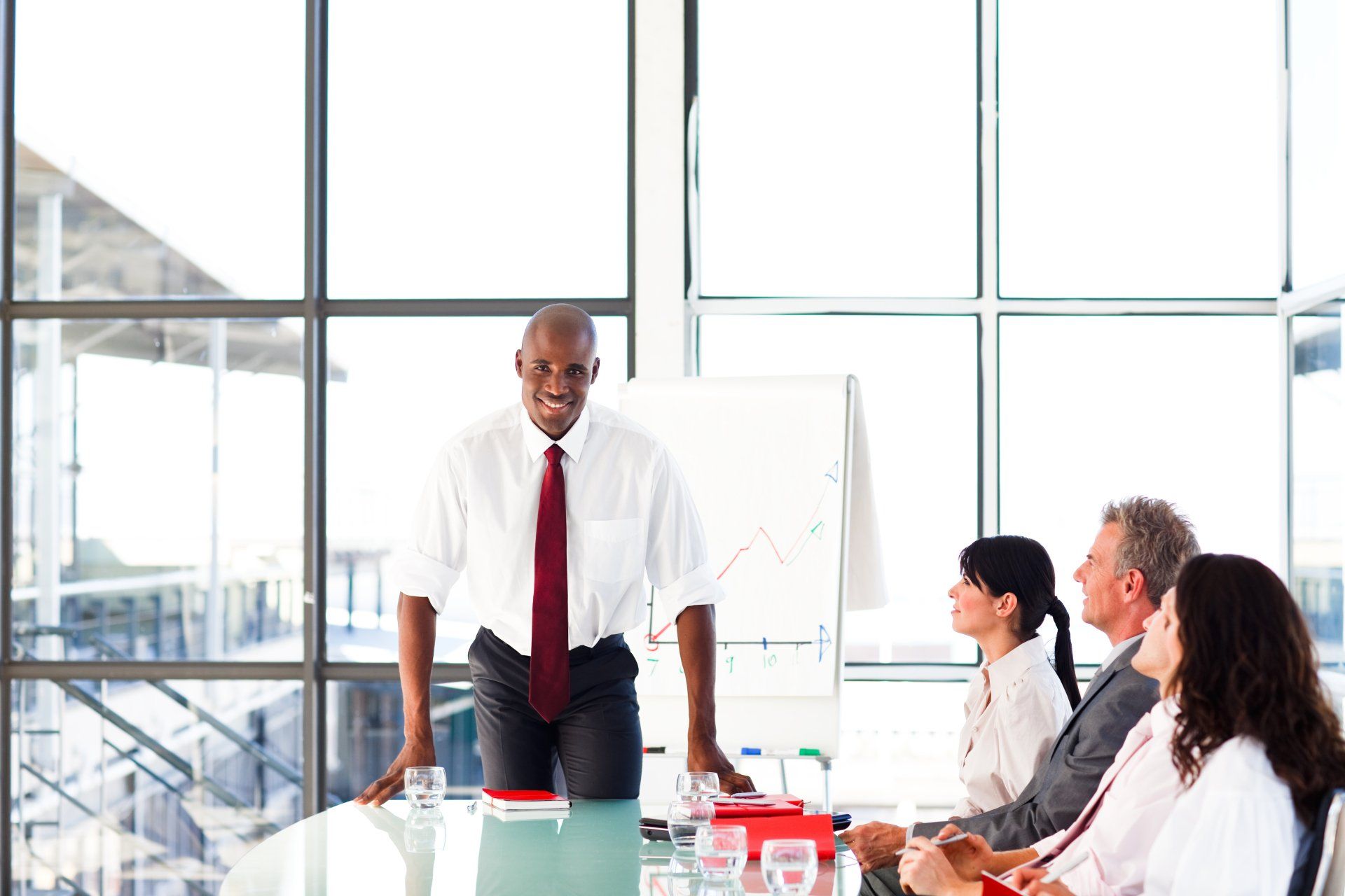 Man giving a presentation at a conference table, smiling. Attendees listen. White shirt, red tie. Bright room with windows.