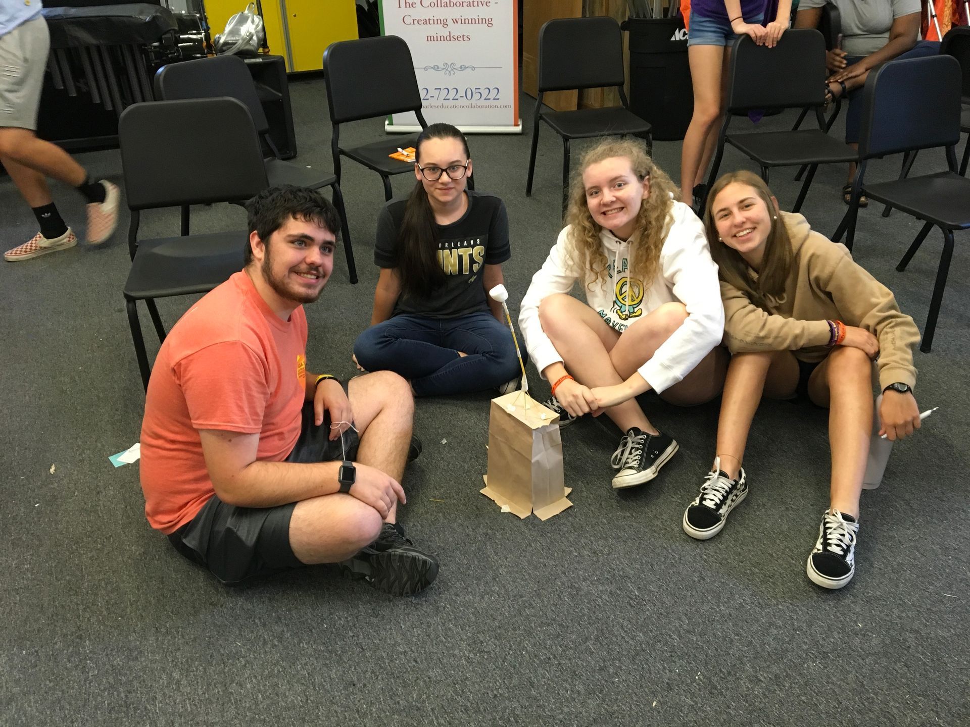Four people sitting around a small paper structure on the floor, smiling.