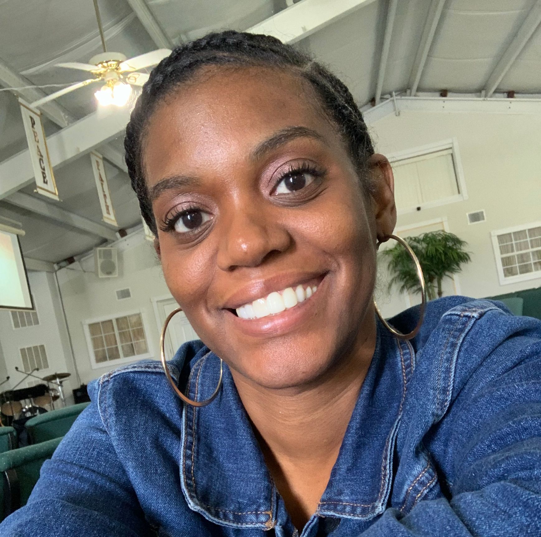 Woman smiles at the camera, wearing a blue jean jacket and large hoop earrings. In a church.