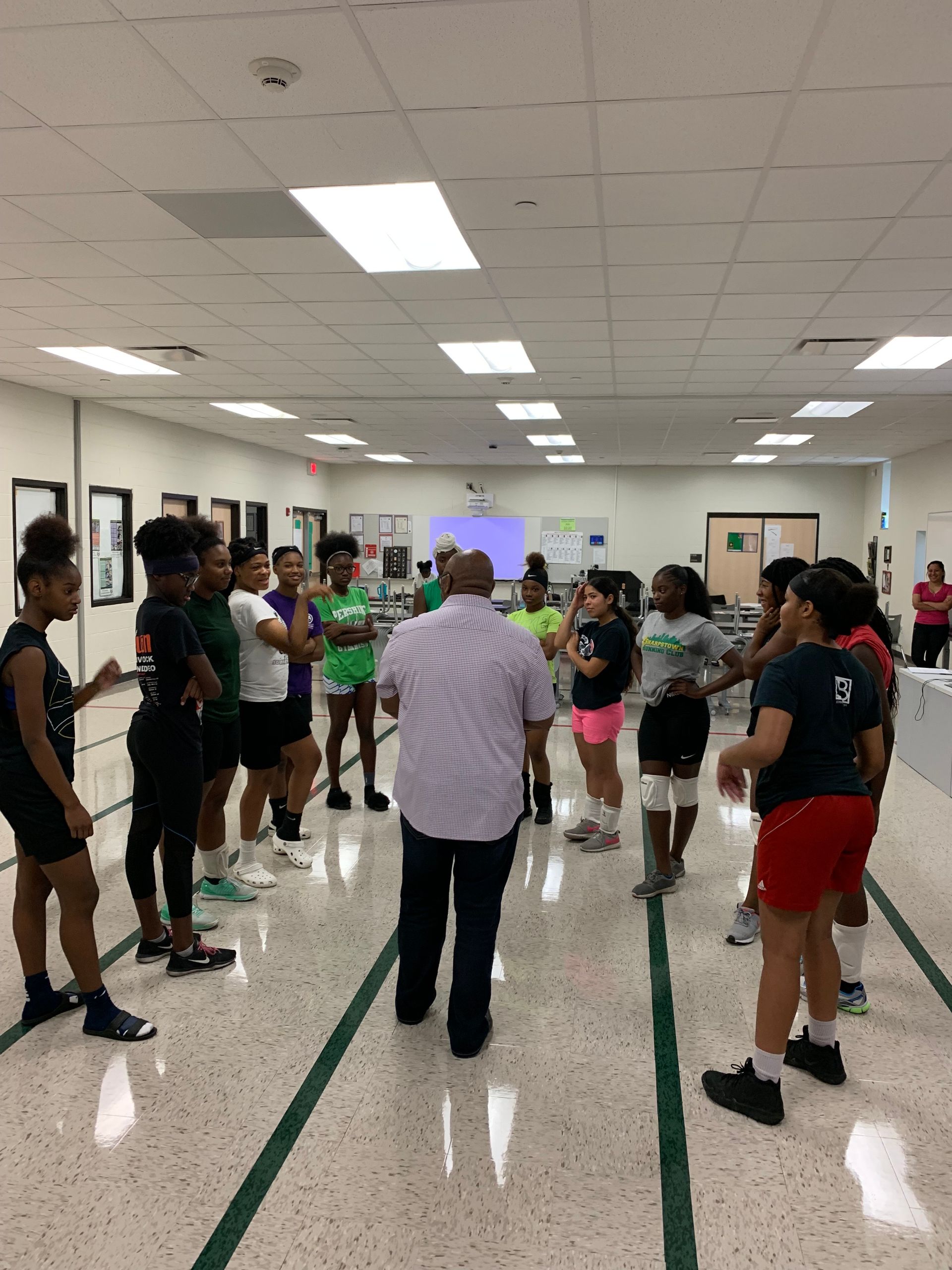 Coach talks to a circle of athletes in a brightly lit room with green and white tile.