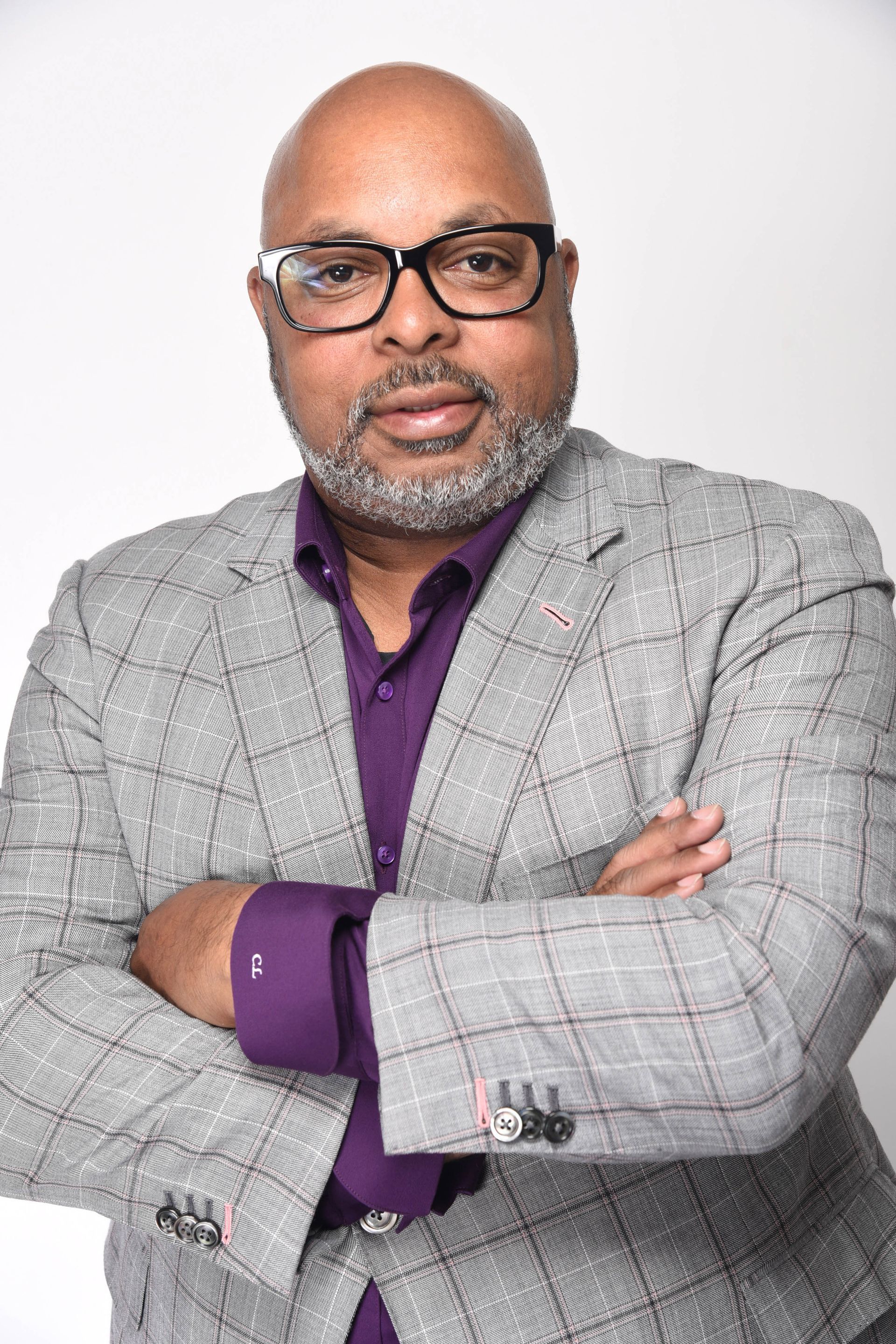 A person wearing glasses, a purple button-down shirt, and a gray checkered blazer, posing with arms crossed.