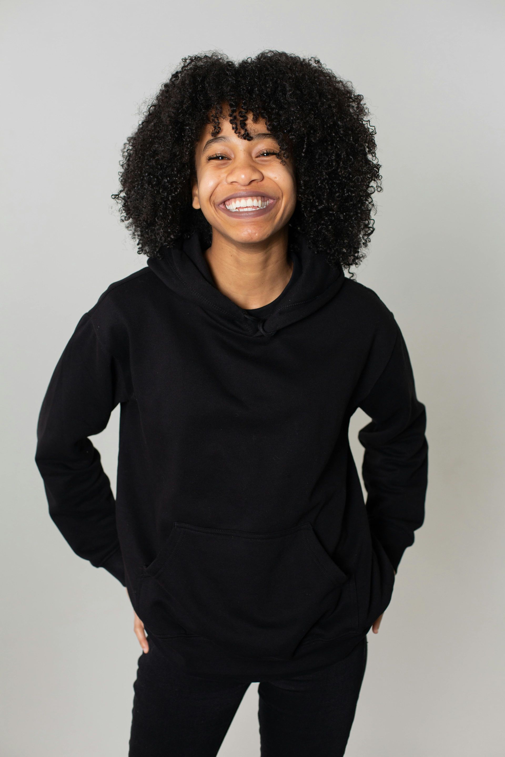 A person with curly hair smiling broadly, wearing a plain black hoodie against a plain gray background.