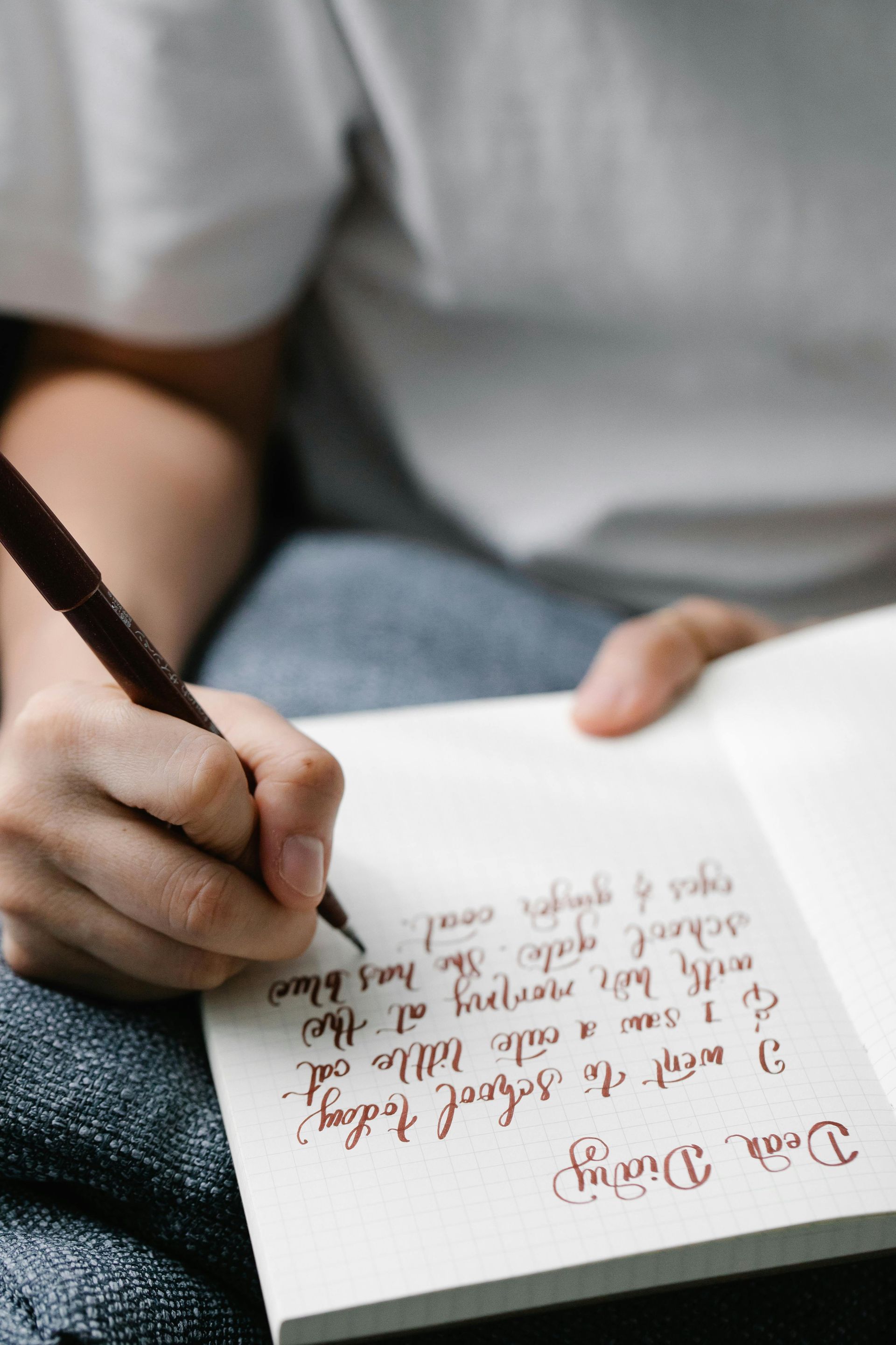 A person in a white shirt writes in a notebook with a dark pen, with handwritten diary text visible on the page.