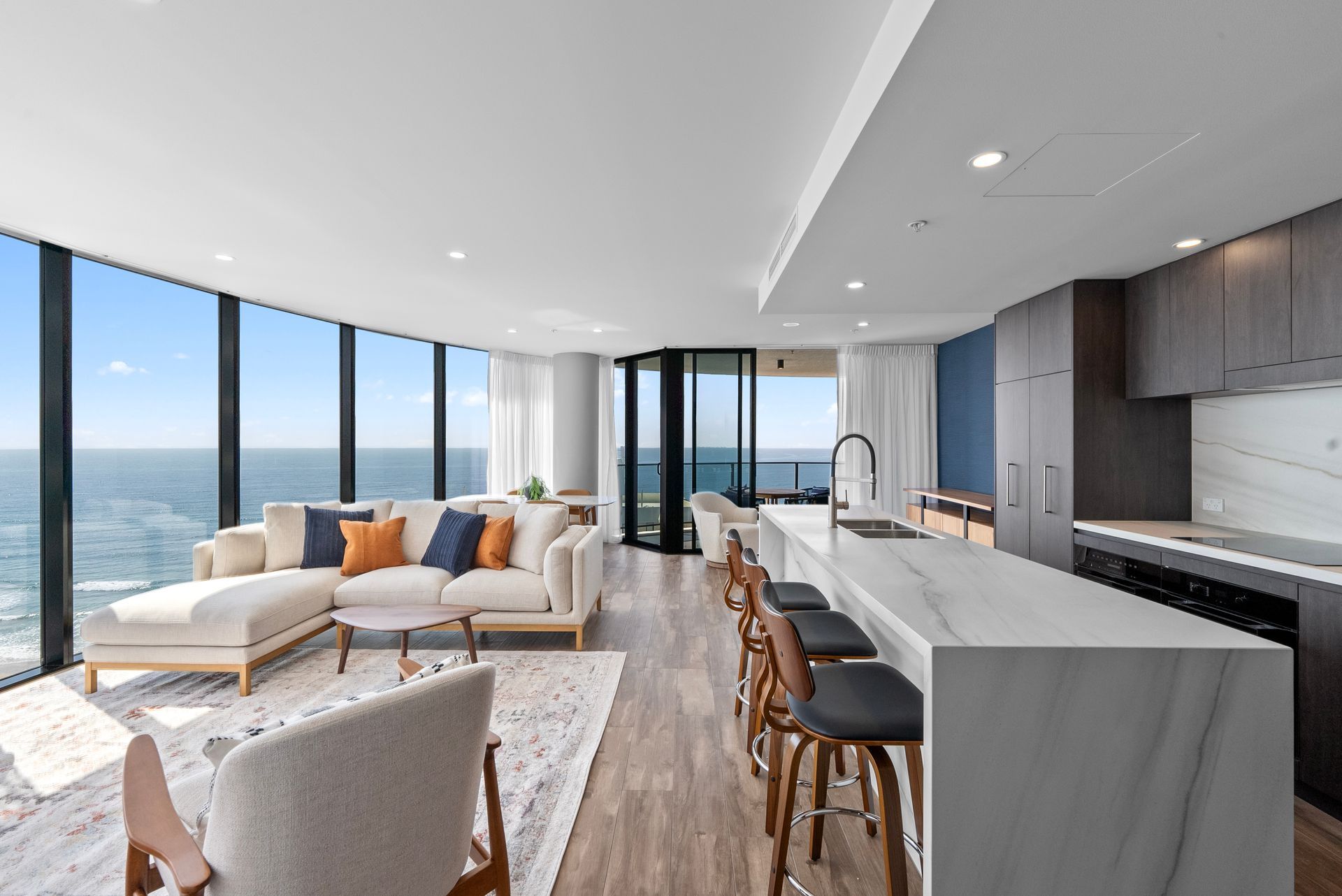 A living room with a couch and chairs and a kitchen with a view of the ocean.