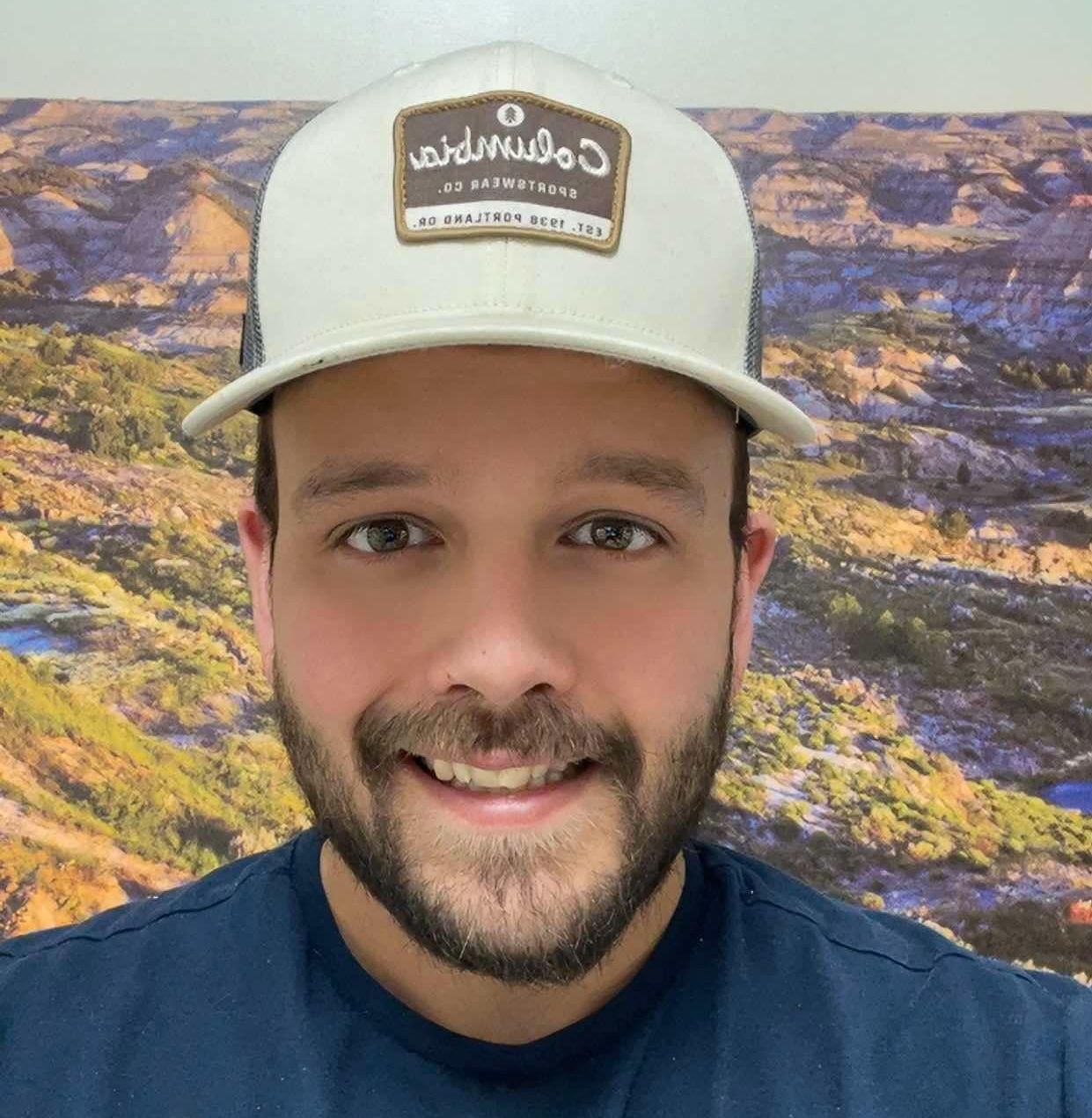 A man with a beard is wearing a white hat and smiling.
