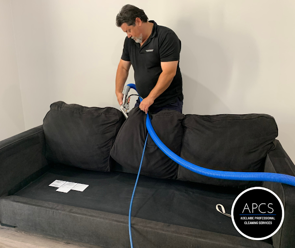 Man cleaning a black couch with a blue hose, in a room with white walls.