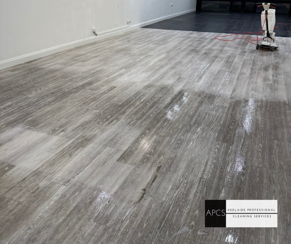 A gray wooden floor being cleaned with a machine in a room with a white wall.