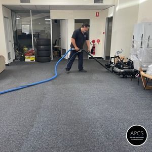 A man cleaning a gray carpet with a blue hose in an office setting.