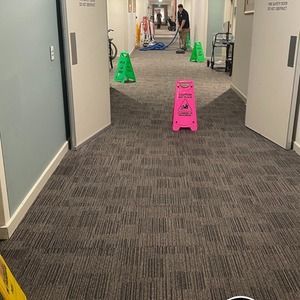 A person vacuums a carpeted hallway, with wet floor signs present.