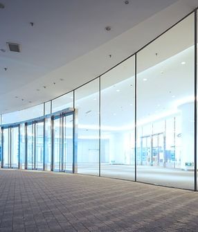 Exterior of a modern building with glass walls and automatic doors, on a tiled walkway.
