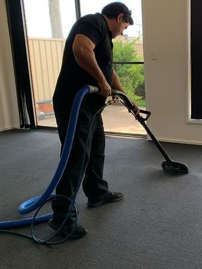 Man in black cleans gray carpet with a vacuum in a room with windows.