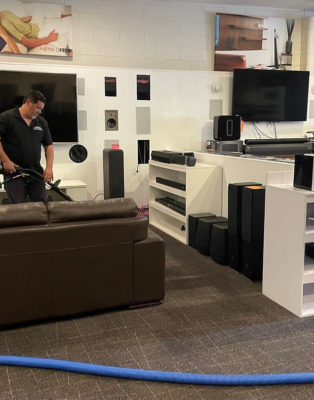 Man vacuuming a brown sofa in a showroom with speakers and TVs on display.