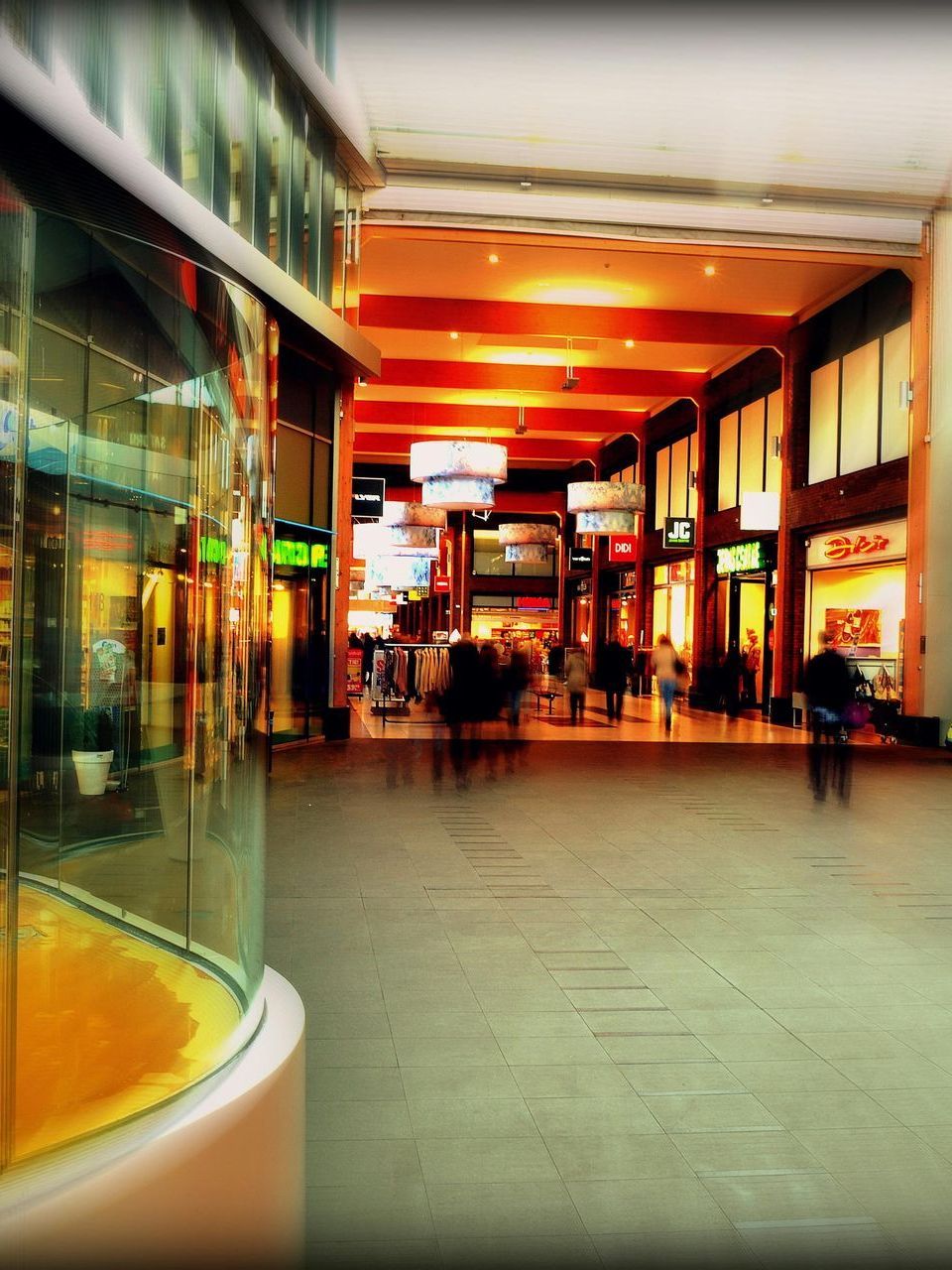 Shopping mall interior, blurred figures walking past stores. Bright lights illuminate the space.