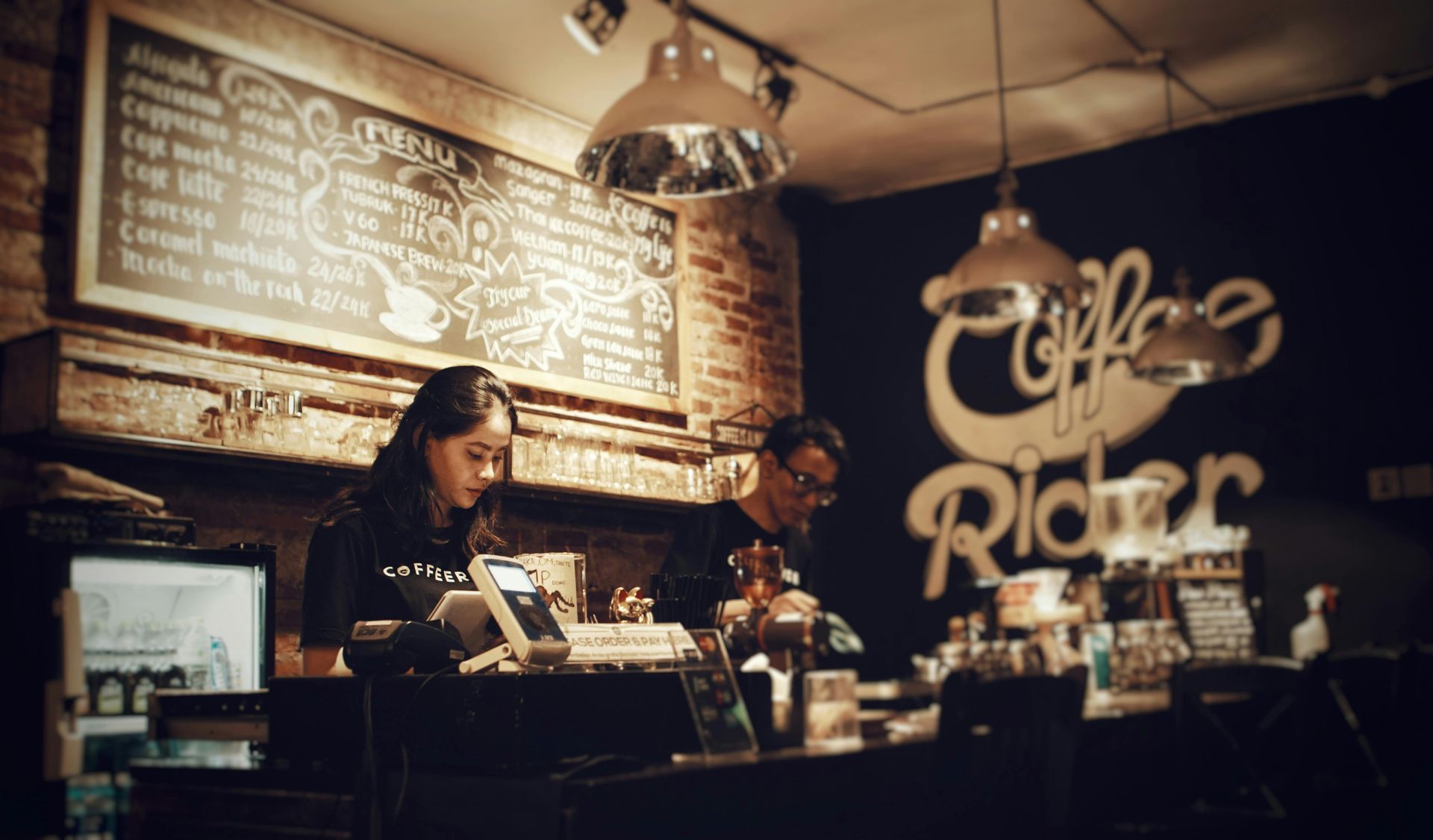Two people working behind the counter at a coffee shop. Dark interior, brick wall, menu, and logo