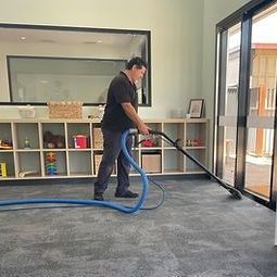 Man vacuums a carpeted floor. He wears a dark shirt, and the setting is a room with shelves.