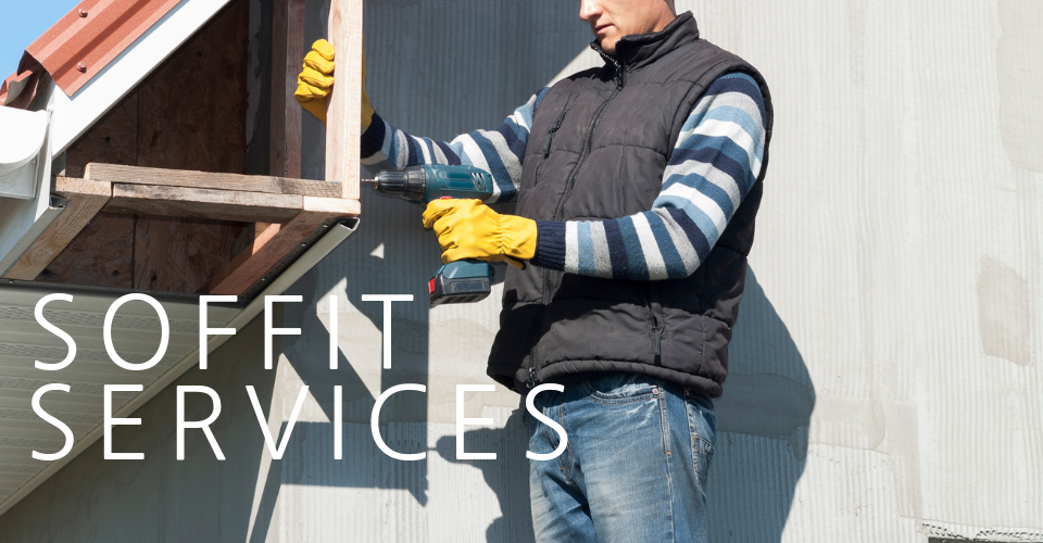 A person wearing a dark vest and yellow gloves uses a power drill to repair a wooden roof soffit.