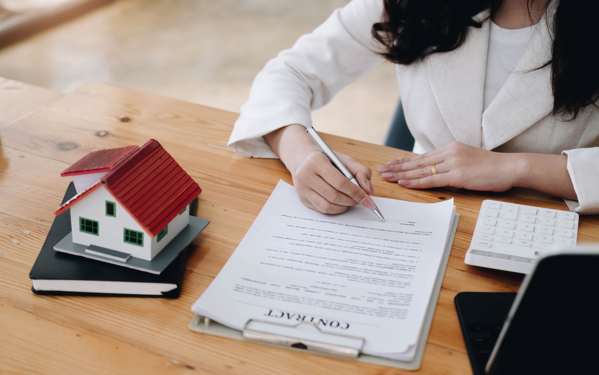 Person signing house loan documents beside a model house on a wooden desk.