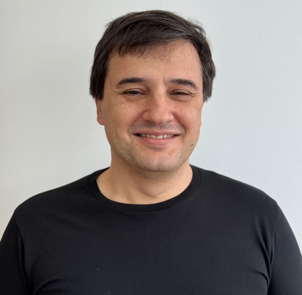 Homem sorrindo, vestindo uma camisa preta, em pé em frente a uma parede branca.