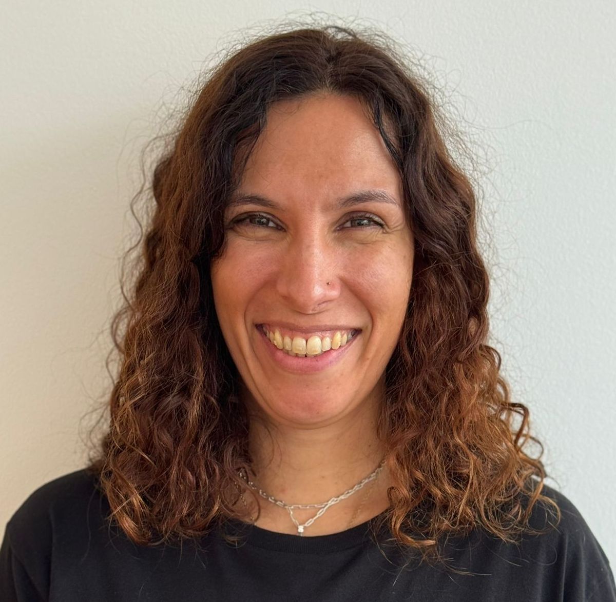 Mulher com cabelos castanhos cacheados sorrindo, vestindo uma camisa preta e um colar prateado, contra uma parede branca.