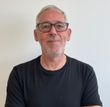 Man with gray hair and glasses in a black t-shirt smiles in front of a white wall.