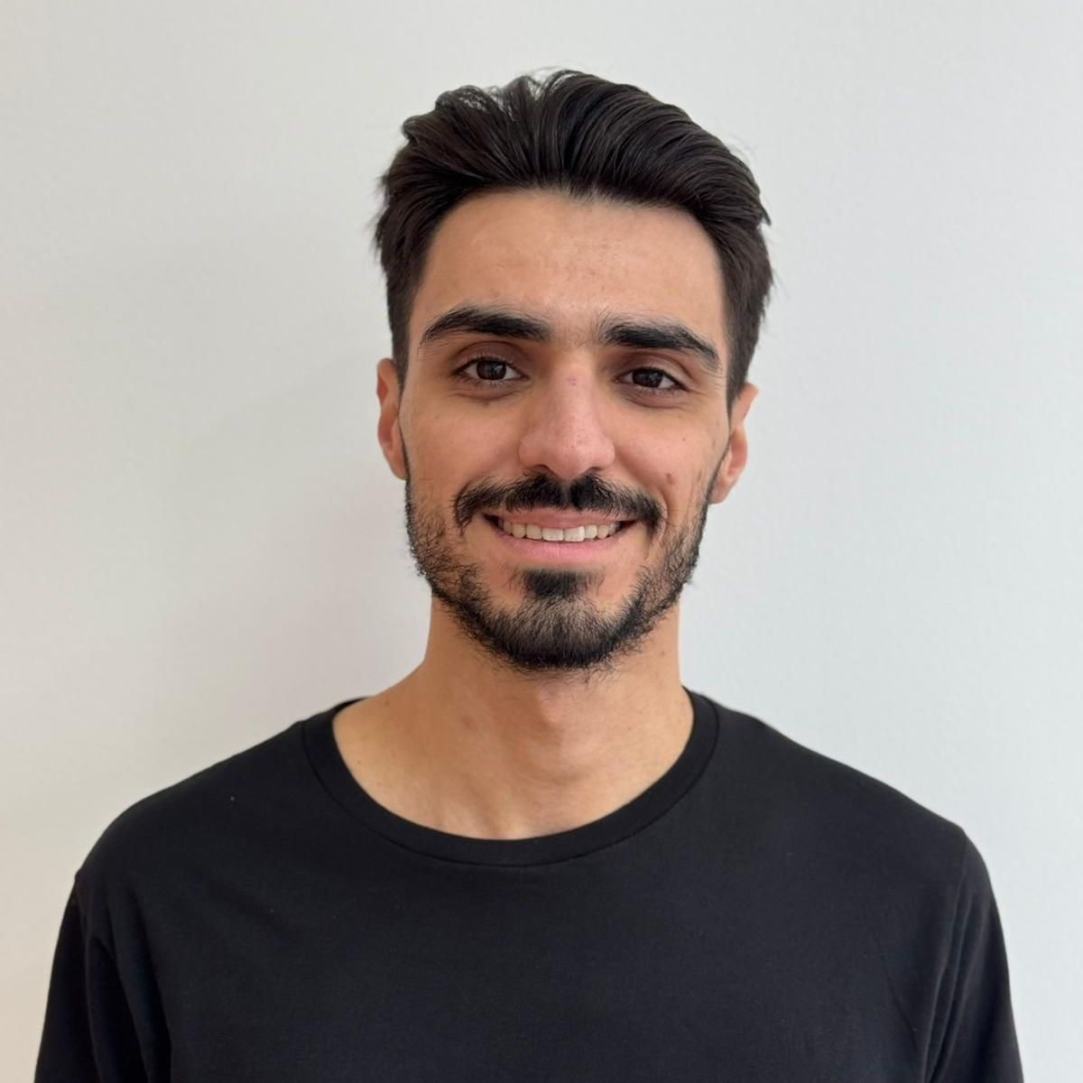 Homem sorridente, cabelo escuro, barba curta, vestindo uma camisa preta, contra uma parede branca.