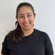 Woman in a black shirt smiles at the camera. Dark hair in a ponytail, white wall backdrop.