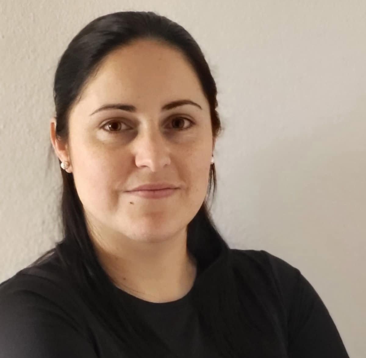Mulher de cabelos escuros, olhando para a câmera com um leve sorriso; vestindo uma camisa preta contra uma parede branca.