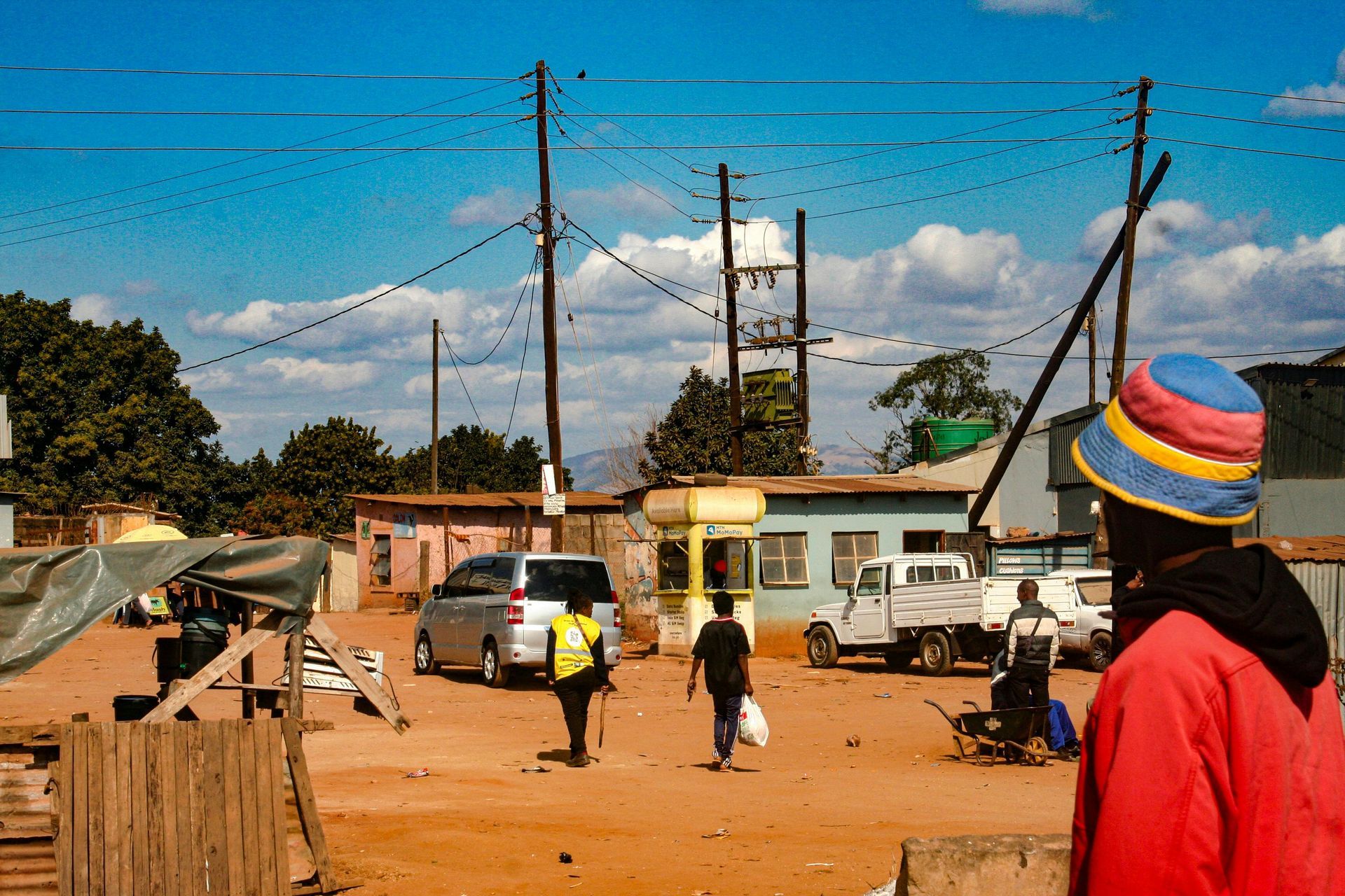 Street scene with power lines and small buildings, people walking; person in red coat and colorful hat in foreground.
