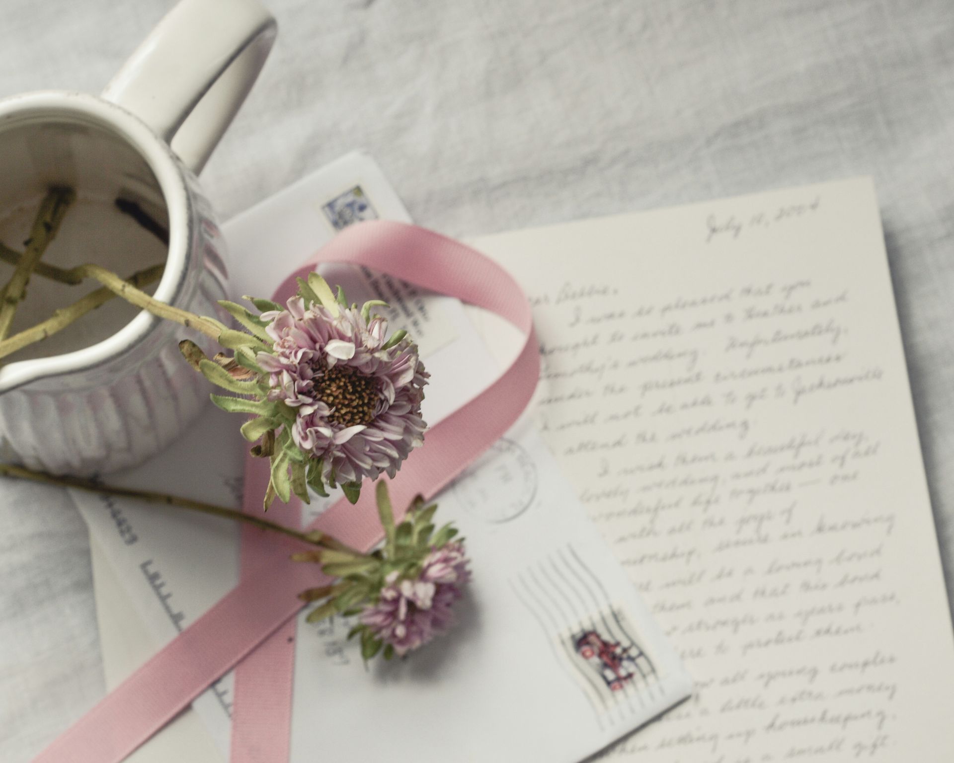 Notebook with handwritten notes, pink ribbon, purple flowers, and a white mug on a soft white surface.