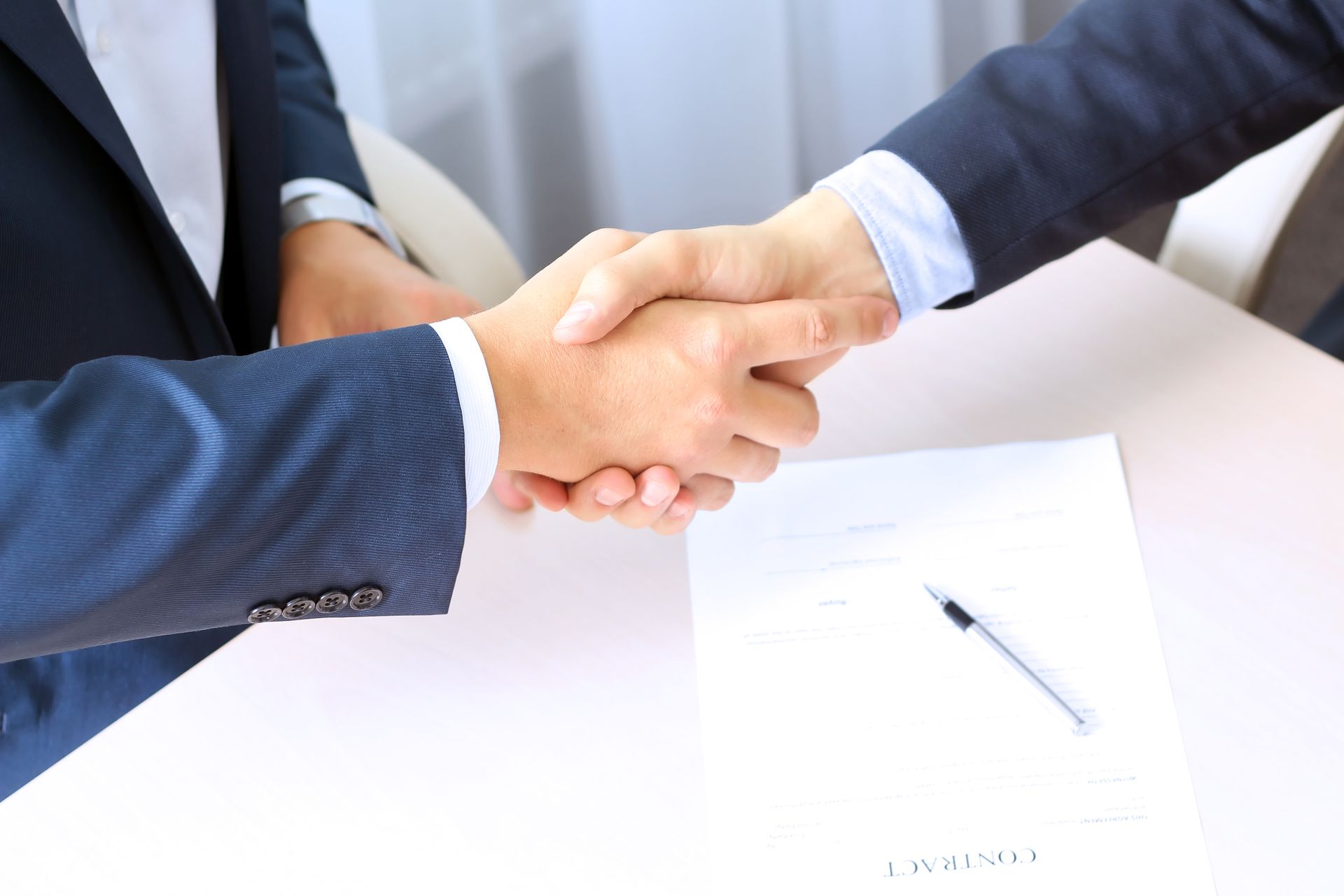 Two people in suits shaking hands over a document on a table.