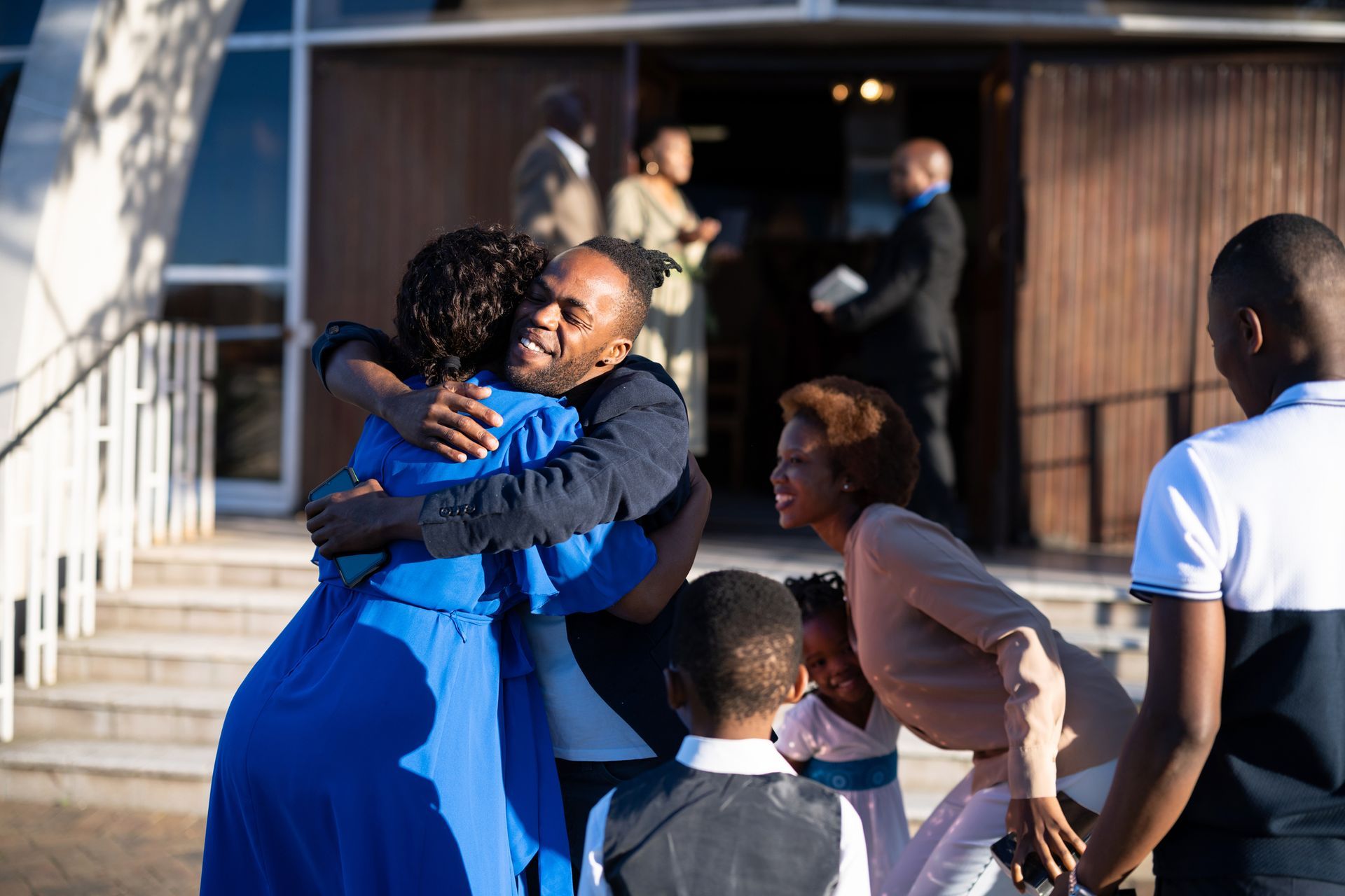 Family embraces outside a church with smiles. People in formal attire, sunny day.