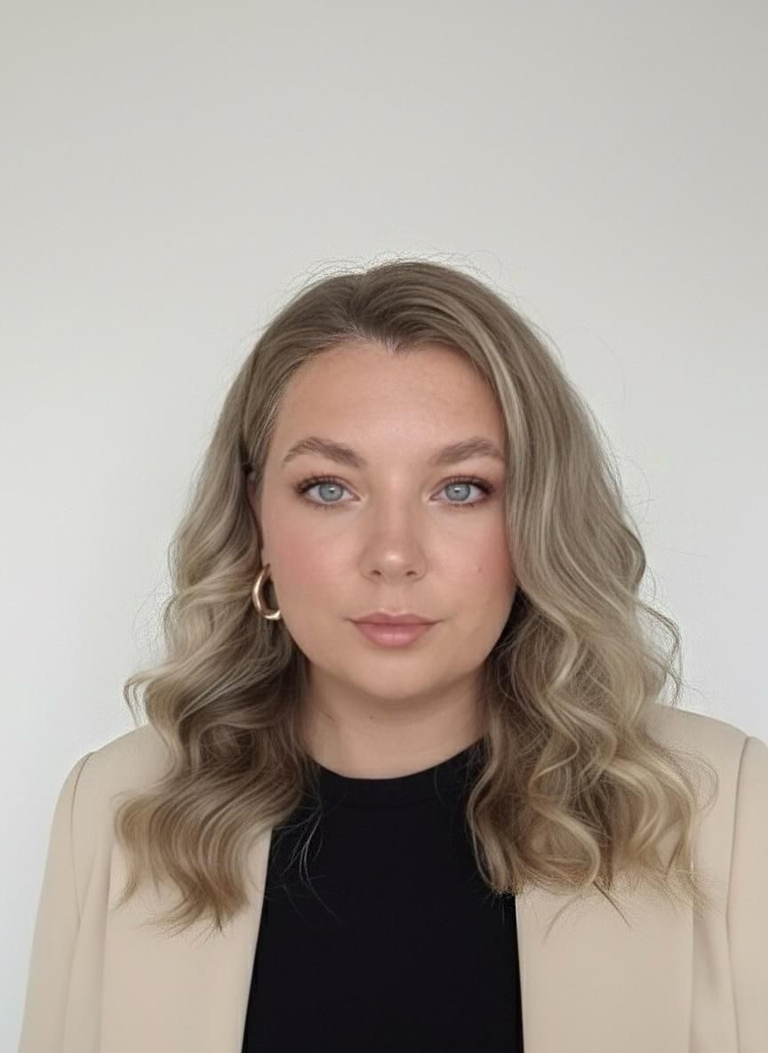 Woman with wavy blonde hair, wearing a black shirt and beige blazer, looking at the camera.