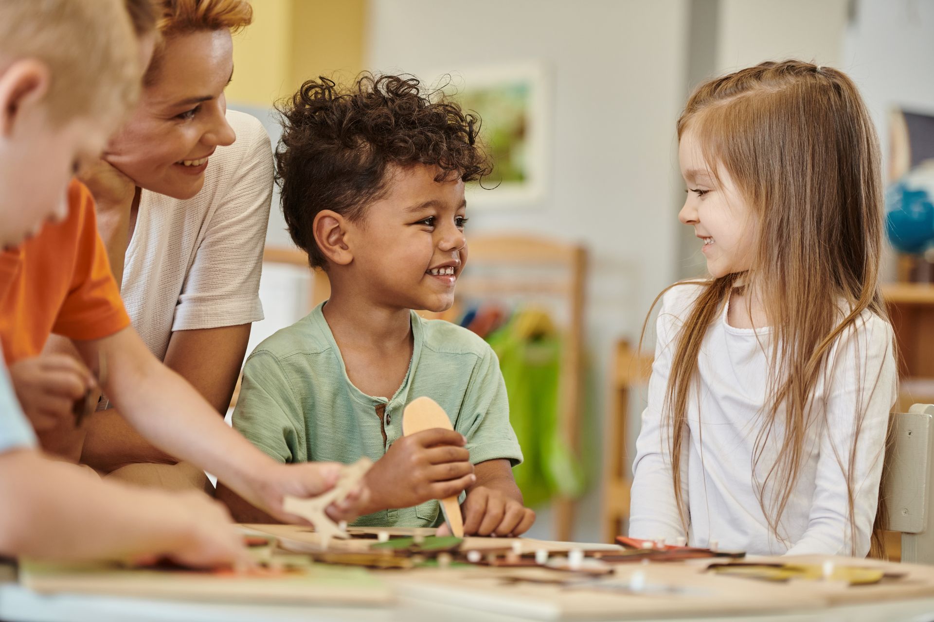 Children and adult teacher at a table, smiling. A boy holds a wooden cutout.