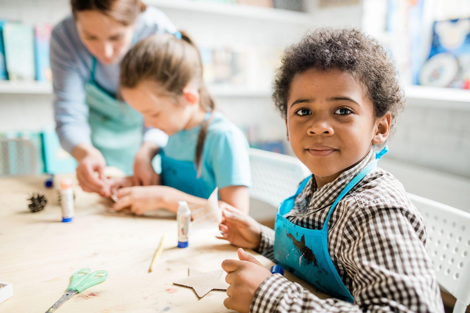 Boy in apron smiles at the camera while crafting at a table with other children and an adult.