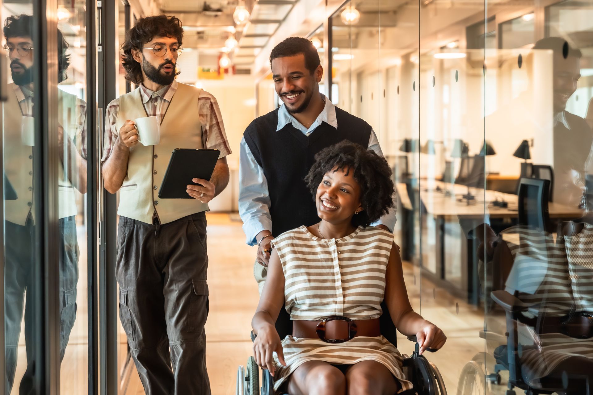 Three people in office hallway; one in wheelchair smiling, being pushed by a person; another person walks holding a tablet.