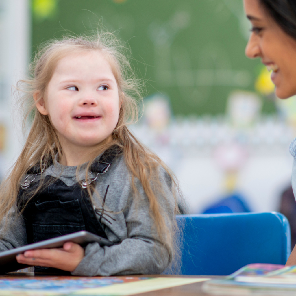 Child smiling at a tablet during a classroom activity with a teacher nearby