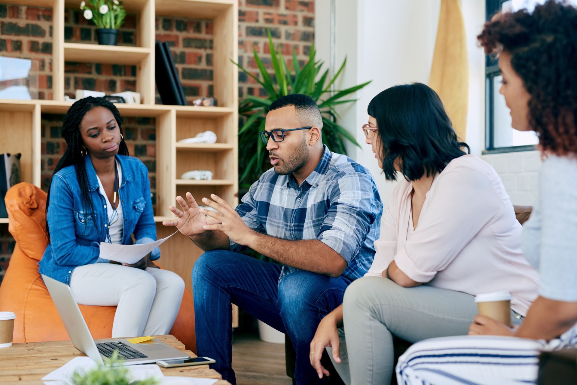 Group of people in discussion, sitting in a casual office setting. Man gestures; others listen, some with papers/laptop.